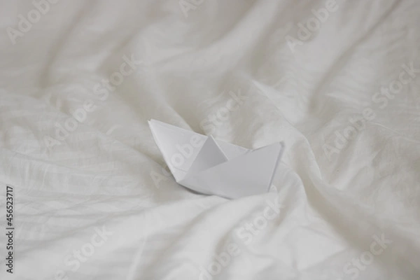 Obraz paper origami boat in bed with waves made with bed sheets, natural white crumpled blanket