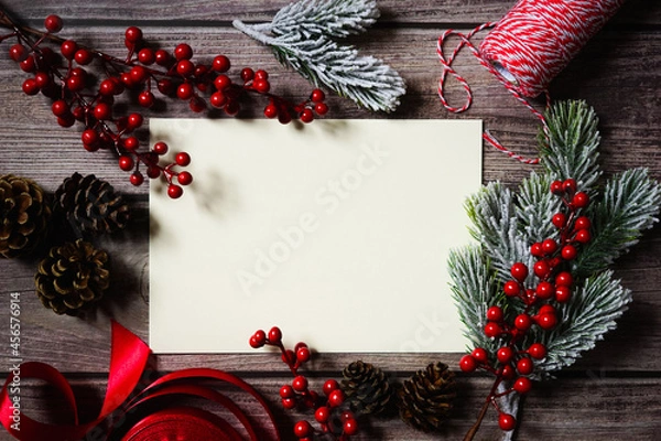 Fototapeta Blank white card surrounded by Christmas decoration on wooden background. Festive holiday greetings. Merry Christmas. Red berries, cones, twine, branches, ribbon. Empty space for text. Copy space.