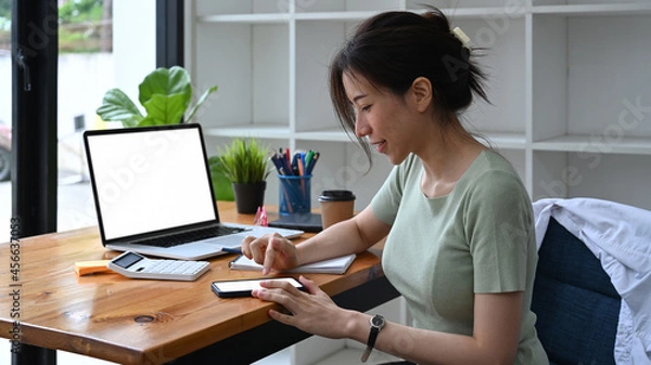Fototapeta Young woman sitting in front of laptop computer and using smart phone.