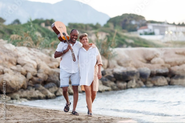 Fototapeta Happy mixed race, middle age couple embracing while walking on the beach holding a guitar. Man holding guitar with woman.
