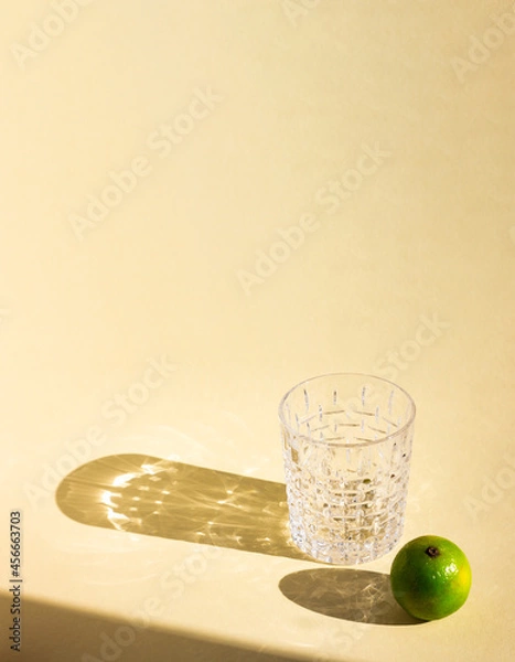 Fototapeta an art creative template of empty crystal glass and lime under hard sunlight, glitter reflection on beige background, long harsh rectangular and oval shadows, copy space