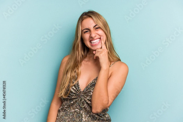 Fototapeta Young caucasian blonde woman isolated on blue background  smiling happy and confident, touching chin with hand.