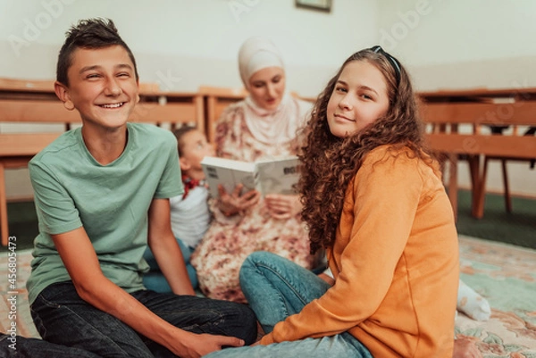 Obraz Female teacher helps school kids to finish they lesson.They sitting all together on floor.Selective focus
