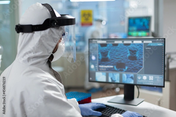 Fototapeta Biologist researcher man wearing coronavirus protective medical equipment while typing covid19 vaccine expertise on computer during biochemistry experiment. Scientist doctor working in clinical lab