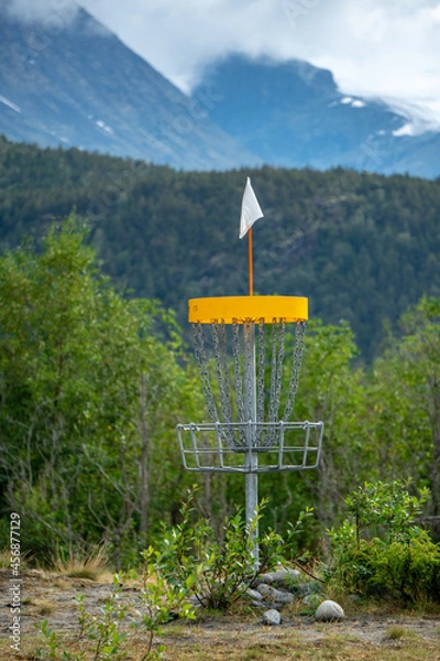 Fototapeta A disc golf hole basket on the ground with the woods and mountains in background during summer time in Norway.