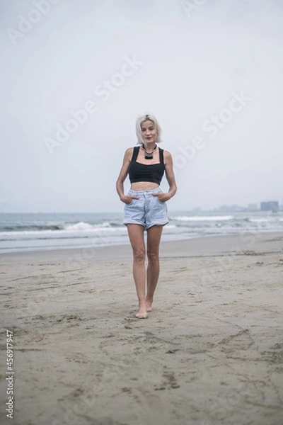 Fototapeta Portrait of pretty caucasian young woman with blond hair, wearing denim shorts, short top and stylish necklace. Walking on the beach. Looks at camera