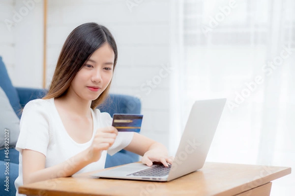 Fototapeta Young asian woman sit on sofa using laptop computer shopping online with credit card buying to internet, happy girl payment with e-business on couch, purchase and payment, business concept.
