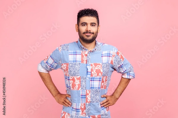 Fototapeta Portrait of happy attractive bearded guy with smile and in casual style blue shirt holding hands on hips, looking at camera with confident expression. Indoor studio shot isolated on pink background.