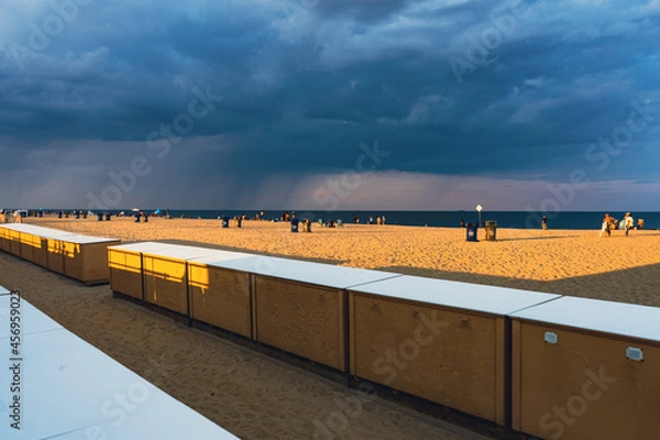 Obraz Beach ocean view with beach lockers in foreground, at sunset. Rainstorm in distance over water. 