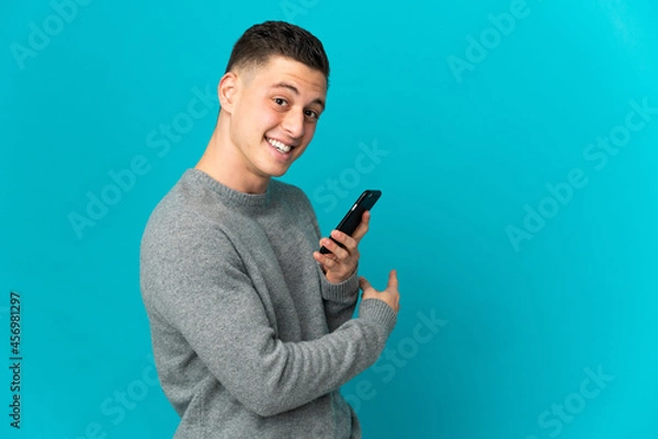 Obraz Young caucasian man isolated on blue background using mobile phone and pointing back