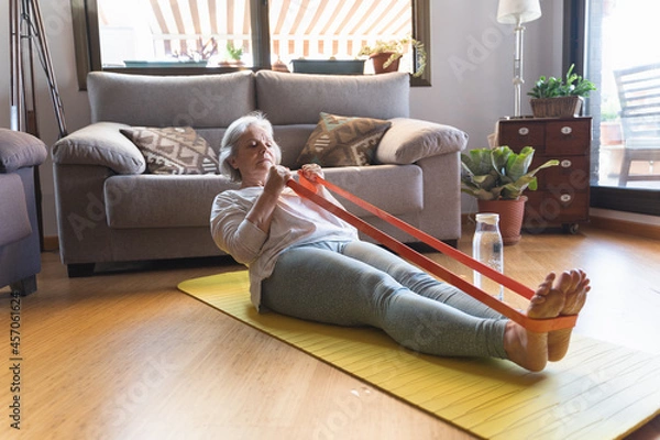 Fototapeta Older woman exercising with an elastic band lying on a mat on the floor of her living room at home