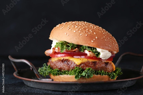 Fototapeta Fast food. Burger with beef, cheese, bacon and vegetables: tomato, onion, cabbage and sauce on a black plate on a black table. Background image, copy space