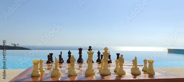 Fototapeta A chessboard, in front of an infinity pool, overlooking the sea, during a sunny, summer day. Chess by the pool.