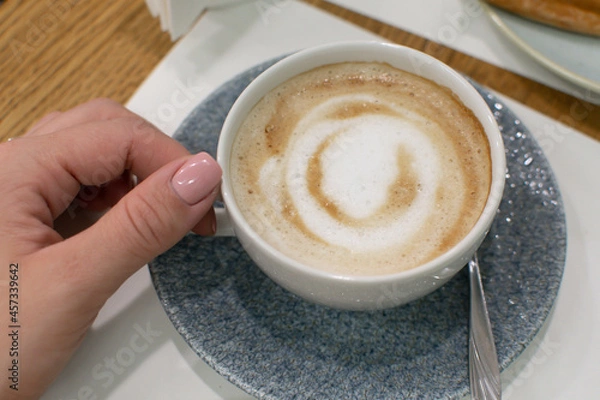 Fototapeta A female hand holds a cup of fresh coffee with milk, a beautiful drawing on top of the foam. Breakfast in a cafe