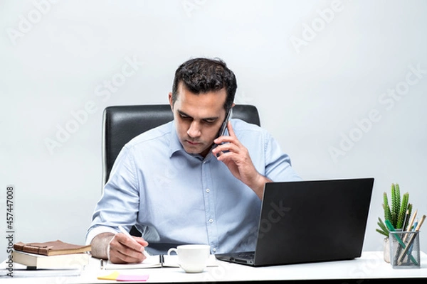 Fototapeta Asian male office worker sitting in front of a laptop computer at a desk and talking on a mobile phone and taking notes at the office.