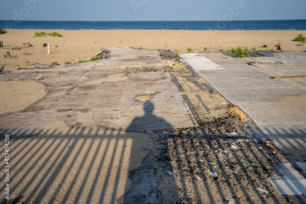 Obraz Shadows of person and fence overlooking entrance to beach from boardwalk. Sand & ocean in distance. 