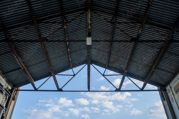 Fototapeta View looking up to industrial building roof from inside, sky & clouds behind.