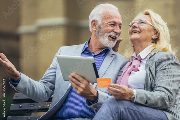 Fototapeta Senior couple sitting in the park on bench and shopping on line with tablet and credit card