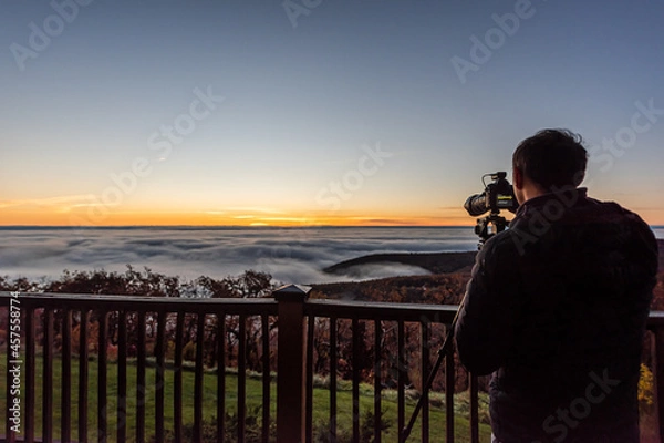 Fototapeta Wintergreen, Virginia ski resort town village with clouds fog mist inversion in Blue Ridge mountains at sunrise and man with camera photographer on apartment balcony