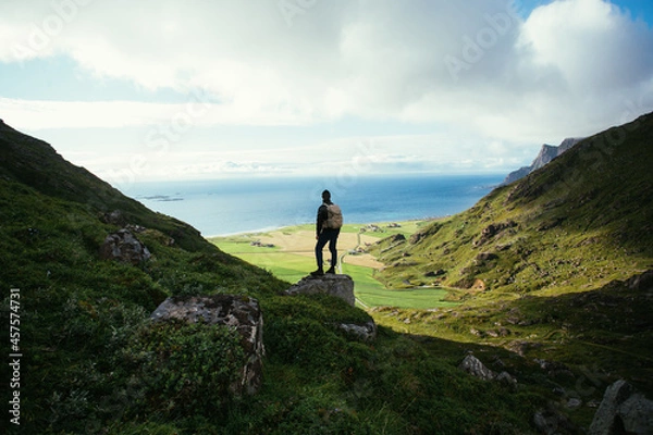Obraz Man with hiking backpack stand on top of mountain, overlook epic views and nordic scandinavian landscape. Modern adventure traveller concept. Young people spend quality time in nature