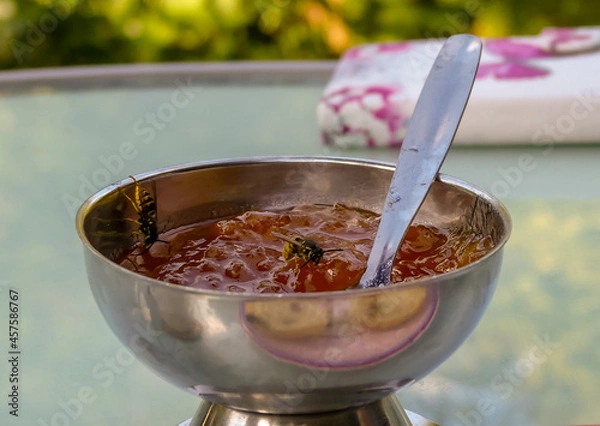 Fototapeta Close up of two common wasps (Vespula vulgaris) feeding on organic plum jam in an open stainless steel bowl on a table top. Visible spoon handle. Outdoor landscape image with space for text. England.