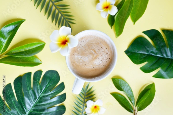 Fototapeta Cup of coffee with tropical leaves on the yellow background. Coffee latte with plumeria in the middle of the table. Coffee break on exotic, tropical island. Happy day, morning. Enjoying vacation.  