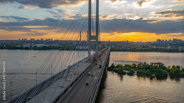 Fototapeta South bridge in Kiev. Sunset over the Dnieper. Thick clouds over the evening city. Evening shot of the bridge. Orange sun at sunset. The rays break through the clouds and are reflected in the river.