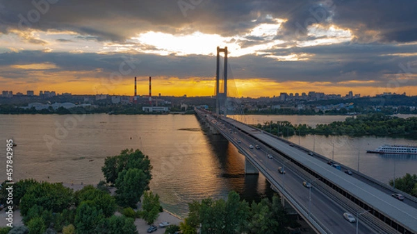 Fototapeta South bridge in Kiev. Sunset over the Dnieper. Thick clouds over the evening city. Evening shot of the bridge. Orange sun at sunset. The rays break through the clouds and are reflected in the river.