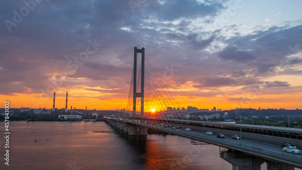 Fototapeta South bridge in Kiev. Sunset over the Dnieper. Thick clouds over the evening city. Evening shot of the bridge. Orange sun at sunset. The rays break through the clouds and are reflected in the river.