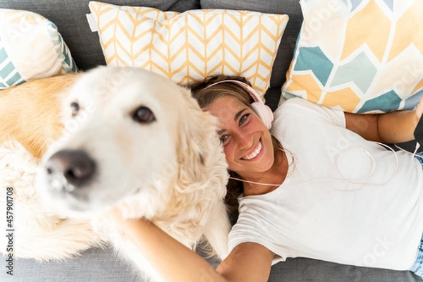 Obraz Selective focus of caucasian blonde woman playing with dog in the couch at home. Top view of caucasian woman listening to music with headphones playing with dog. People and dogs lifestyles.