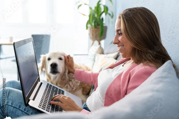 Fototapeta Cheerful blonde woman working on computer with pet on the couch at home. Horizontal side view of caucasian woman playing with her dog indoors. Technology, animals and people lifestyle.