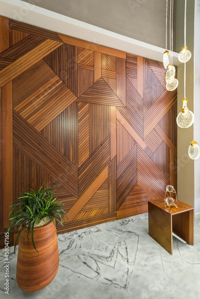 Fototapeta Angled view of hall with wood cladding wall decorated with geometrical patterns, modern glass LED tall chandelier, tall planter with green bushes, and white marble floor in modern house