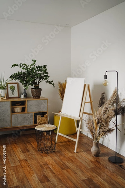 Fototapeta Home art studio interior. Easel with white canvas and brushes in the interior of the room. Concept of hobby drawing, canvas mockup.