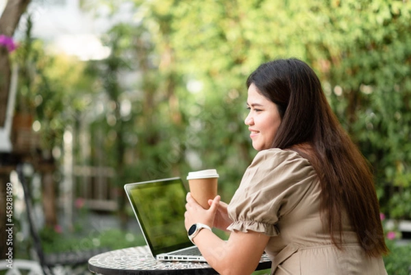 Fototapeta Rear view of Asian woman is using her laptop to check email and work via the internet with a coffee next to her ,Working from home concept.