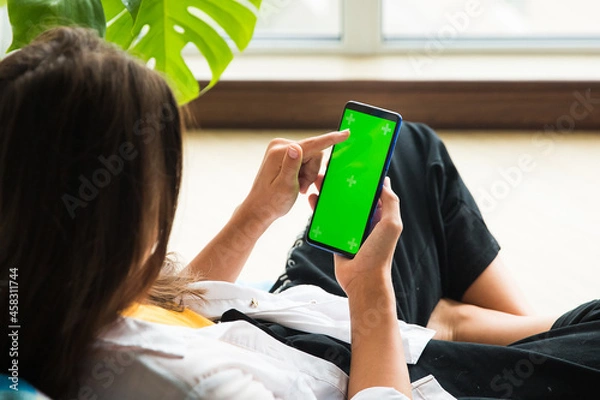Obraz a vertical smartphone with a chromakey screen in the hand of a girl who is sitting by the window
