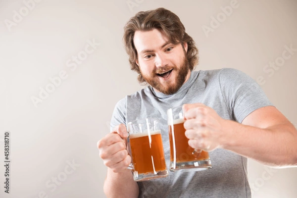 Fototapeta Portrait of a happy young bearded man holding two beer mugs isolated over light gray background