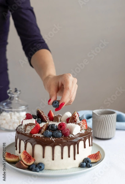 Fototapeta Woman decorating birthday cake with fresh berries and chocolate ganache. delicious homemade sweet baked dessert. gray wall background. vertical image