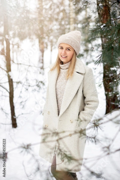 Fototapeta A beautiful girl and smiles in a coat and a hat stands in a snowy forest