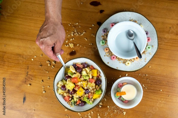 Obraz man mixing fruit salad with silver spoon, in white bowl, decorated with colored cereals, and yogurt and cereal spread around on wooden table in photo taken from above.