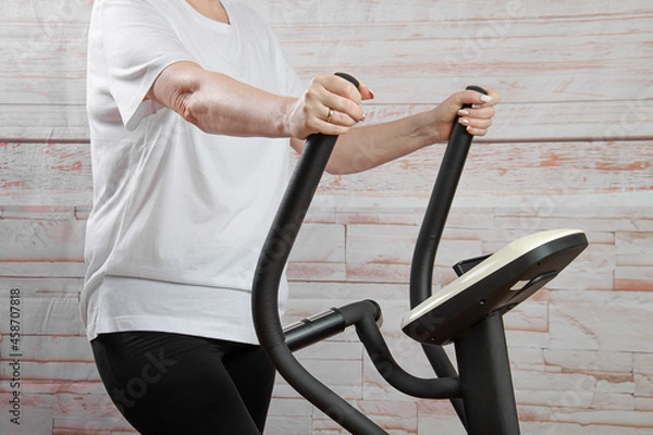 Fototapeta A young middle-aged woman goes in for sports on an elliptical exercise machine.