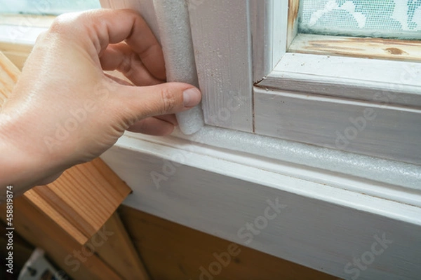 Fototapeta Woman hand insulating old windows to prevent warmth heat leak and drafts, preparing house for winter and cold weather