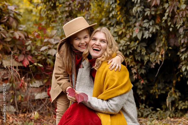 Obraz A young adult blonde mother with a 7-year-old daughter on a walk in the autumn park hug and laugh
