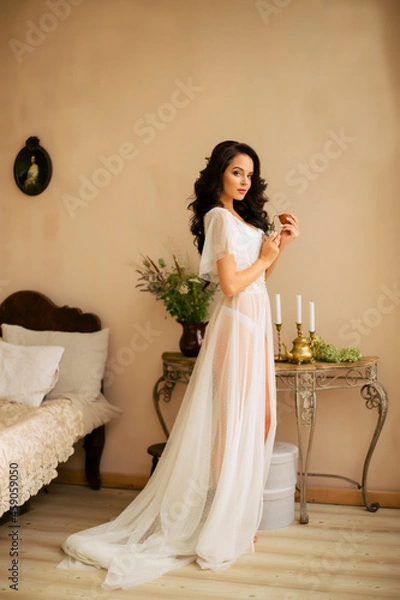 Fototapeta Young beautiful bride stands in a white dress holding perfume in her hands. Brunette of European appearance. Model looks at the camera with her face.