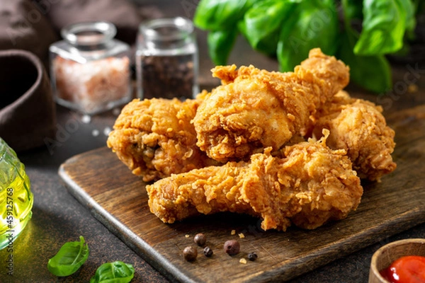 Obraz Fried crispy chicken legs in breadcrumbs on a wooden serving board on a dark background. Fast food from the drumstick close-up