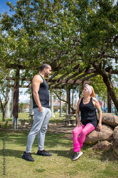 Obraz brown athletic boy talking to blonde lady wearing pink pants and black t-shirt, sitting on rock in park on sunny day.

