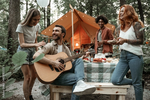 Fototapeta Group of young friends traveling in glamping in the forest having fun playing guitar and mbira kalimba roasting sausages sitting at dinner table near tent during summer vacation laughing hanging out