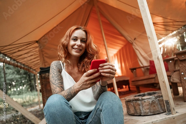 Fototapeta Red-haired smiling girl sits next to the tent and chatting messaging streaming on the smartphone, posting, liking in social media. Travel outside city in the woods. Camping.