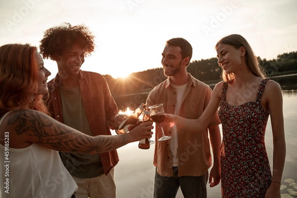 Fototapeta Group of friends having fun on picnic near a lake or river, drinking wine, beer, cider clinking glasses while saying a toast. Smiling young people partying outdoors during sunset in countryside.