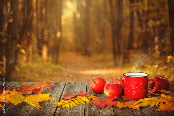 Fototapeta Red mug of tea with apples and leaves on a wooden table and autumn forest background.