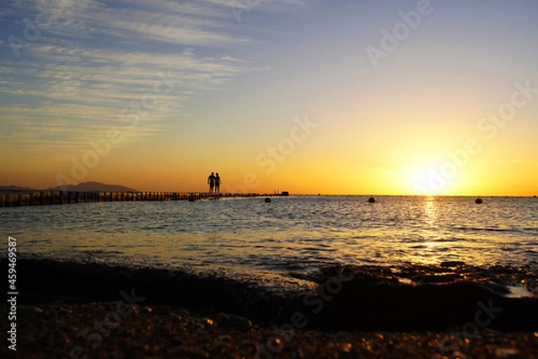 Obraz Colorful sunset by the ocean, two people walk across the bridge, two silhouettes             